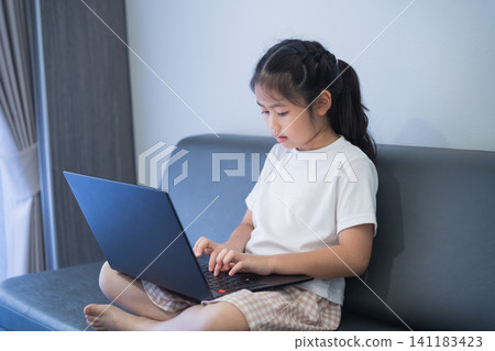 Young girl sitting on a couch using a laptop with focused expression, natural light coming through the window, casual clothing, indoors home environment Young girl sitting on a couch using a laptop with focused expression, natural light coming through the window, casual clothing, indoors home environment 141183423