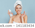 Smiling woman with towel, holding skincare bottle against light blue background 141183434