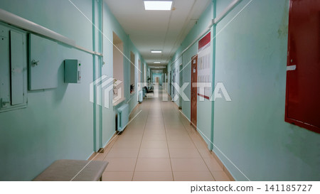 Long empty hospital corridor with pale green walls, beige tiled floor, ceiling light panels, handrails, radiators, and closed wooden doors receding toward distant vanishing point Long empty hospital corridor with pale green walls, beige tiled floor, ceiling light panels, handrails, radiators, and closed wooden doors receding toward distant vanishing point 141185727