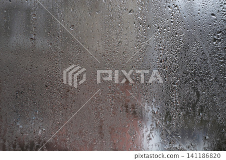 Wet glass with dense raindrops and vertical water lines. glass creating abstract background with soft blurred house exterior. dampness window texture and condensation Wet glass with dense raindrops and vertical water lines. glass creating abstract background with soft blurred house exterior. dampness window texture and condensation 141186820
