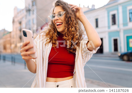 Optimistic young woman looking at camera with smile while taking selfie on city street. Optimistic young woman looking at camera with smile while taking selfie on city street. 141187026