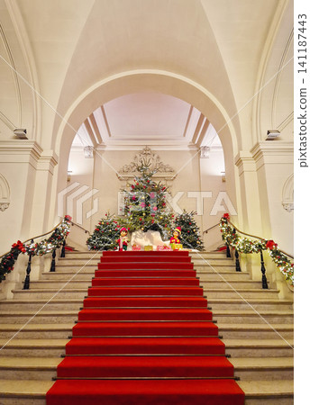 A grand stone staircase with a red carpet leads up to a Christmas tree display in Asnieres-sur-Seine city hall. Festive garlands, elf figures and wrapped gifts under magnificent neoclassical arches A grand stone staircase with a red carpet leads up to a Christmas tree display in Asnieres-sur-Seine city hall. Festive garlands, elf figures and wrapped gifts under magnificent neoclassical arches 141187443