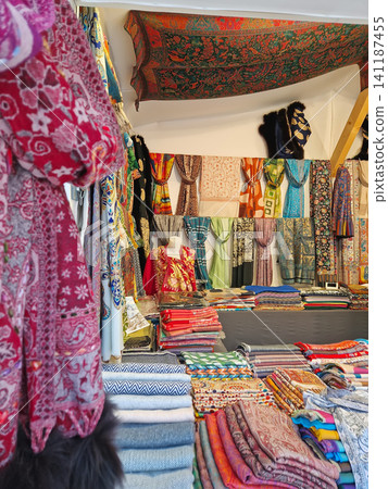 Paris, France - December 10, 2025: vibrant market stall is filled with colorful, patterned scarves, shawls, and textiles. Stacks of folded fabrics of rich, intricate craftsmanship rest on the counter Paris, France - December 10, 2025: vibrant market stall is filled with colorful, patterned scarves, shawls, and textiles. Stacks of folded fabrics of rich, intricate craftsmanship rest on the counter 141187455