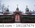 Snowflakes falls gently over an Orthodox monastery, dusting its domes and golden crosses in white snow, while festive lights twinkle in the twilight. Serene winter scene capturing the spiritual beauty 141187479