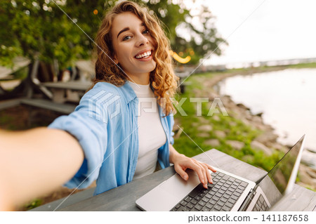 Happy woman sitting at a table outdoors taking a selfie, working on a laptop with view of the lake. Happy woman sitting at a table outdoors taking a selfie, working on a laptop with view of the lake. 141187658