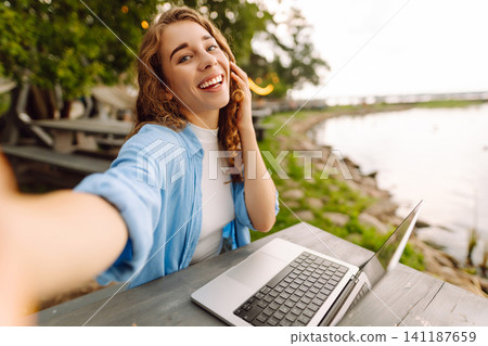 Happy woman sitting at a table outdoors taking a selfie, working on a laptop with view of the lake. Happy woman sitting at a table outdoors taking a selfie, working on a laptop with view of the lake. 141187659