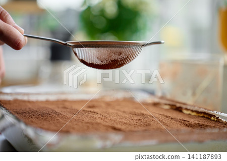 Hand holds sieve with cocoa powder over tiramisu dessert in glass dish Hand holds sieve with cocoa powder over tiramisu dessert in glass dish 141187893