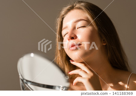Young woman reflected in the mirror in soft evening light in a cozy room. Beauty and care concept. 141188369