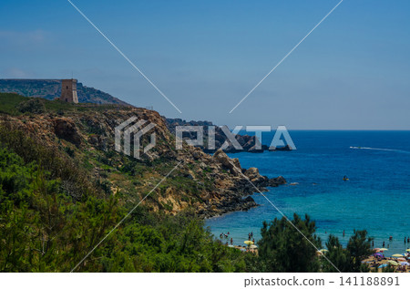 Beautiful Malta coastline with old stone watchtower on rugged cliffs, green vegetation and beach umbrellas beside crystal clear turquoise Mediterranean sea. Beautiful Malta coastline with old stone watchtower on rugged cliffs, green vegetation and beach umbrellas beside crystal clear turquoise Mediterranean sea. 141188891