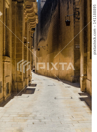 Narrow empty medieval street with golden limestone buildings and old lanterns in Mdina, historic Malta town alleyway Narrow empty medieval street with golden limestone buildings and old lanterns in Mdina, historic Malta town alleyway 141188895