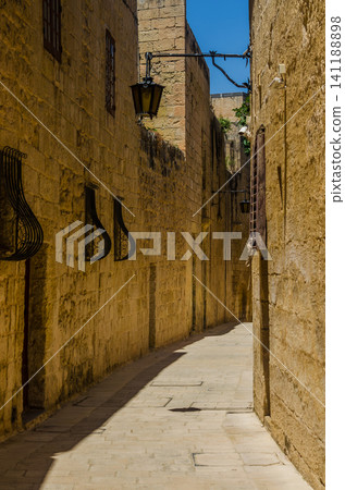 Empty narrow medieval street with golden limestone walls, iron lanterns and window grilles in historic Mdina, Malta Empty narrow medieval street with golden limestone walls, iron lanterns and window grilles in historic Mdina, Malta 141188898