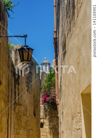 Narrow medieval alley with old lantern, pink bougainvillea and church dome in golden limestone street in Mdina, Malta Narrow medieval alley with old lantern, pink bougainvillea and church dome in golden limestone street in Mdina, Malta 141188901