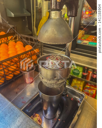 Traditional manual citrus press extracting fresh pomegranate juice at street market stall with crate of ripe oranges nearby Traditional manual citrus press extracting fresh pomegranate juice at street market stall with crate of ripe oranges nearby 141188904