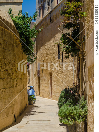 Narrow sunlit Mediterranean alleyway with rustic limestone walls, green plants, and a parked blue scooter in an old historic town. Narrow sunlit Mediterranean alleyway with rustic limestone walls, green plants, and a parked blue scooter in an old historic town. 141188912