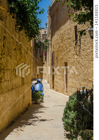 Narrow sunlit Mediterranean alleyway with rustic limestone walls, green plants, and a parked blue scooter in an old historic town. Narrow sunlit Mediterranean alleyway with rustic limestone walls, green plants, and a parked blue scooter in an old historic town. 141188915