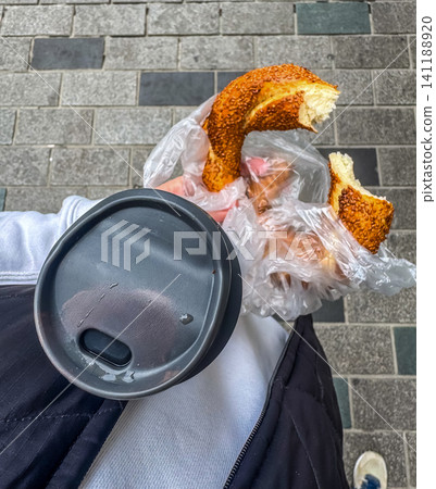 Person holding sesame bread ring simit and takeaway coffee cup while walking on cobblestone street point of view first person Person holding sesame bread ring simit and takeaway coffee cup while walking on cobblestone street point of view first person 141188920