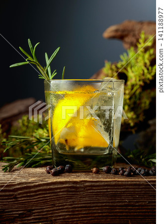 Gin and tonic cocktail with ice, lemon, juniper berries and rosemary sprigs sits on an old weathered board. 141188977