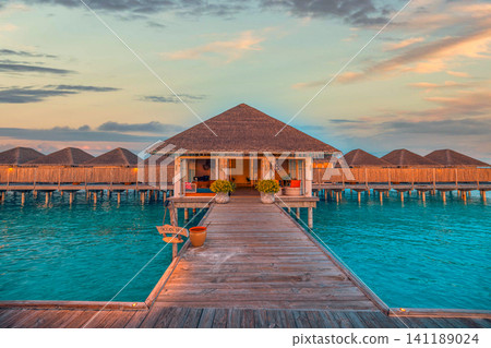Overwater bungalows and wooden jetty at colorful sunset in Maldives resort. Luxury water villas above turquoise lagoon under dramatic evening sky, warm lights on pier. Iconic honeymoon destination Overwater bungalows and wooden jetty at colorful sunset in Maldives resort. Luxury water villas above turquoise lagoon under dramatic evening sky, warm lights on pier. Iconic honeymoon destination 141189024