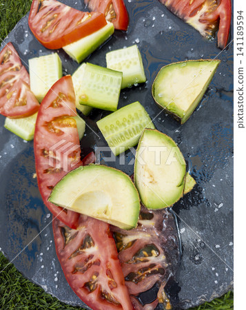 Fresh slices of red tomatoes, green cucumbers, and ripe avocado are arranged on a black slate platter. Sea salt adds flavor to this summer salad. Fresh slices of red tomatoes, green cucumbers, and ripe avocado are arranged on a black slate platter. Sea salt adds flavor to this summer salad. 141189594