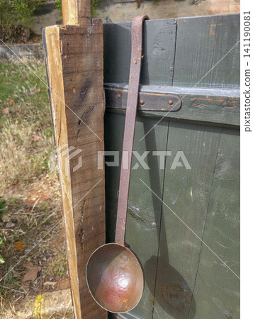 A rustic metal ladle is hanging on a dark green wooden fence panel. The setting is outdoors with some grass and dirt visible. The ladle shows signs of wear and age. A rustic metal ladle is hanging on a dark green wooden fence panel. The setting is outdoors with some grass and dirt visible. The ladle shows signs of wear and age. 141190081