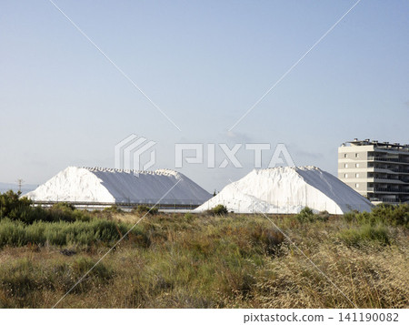 Piles of white sea salt sit at a processing plant beside a modern building in a residential area. Wild fields fill the space around the site, showing a mixture of nature and industry. Piles of white sea salt sit at a processing plant beside a modern building in a residential area. Wild fields fill the space around the site, showing a mixture of nature and industry. 141190082