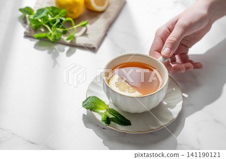 Hand holding a cup of tea with lemon, ginger and mint on a marble background Hand holding a cup of tea with lemon, ginger and mint on a marble background 141190121