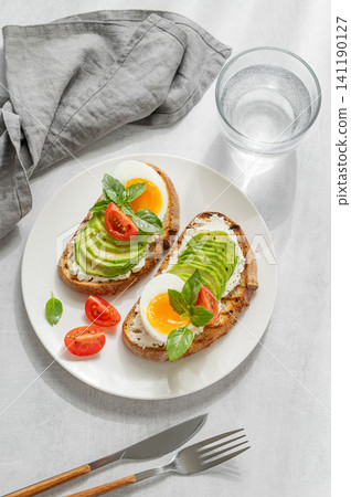 Two avocado toasts with egg, cream cheese, tomatoes, and basil on a concrete background Two avocado toasts with egg, cream cheese, tomatoes, and basil on a concrete background 141190127