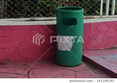 Green cylindrical public trash bin set against a pink hexagonal textured wall 141190245