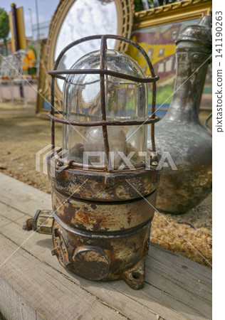 An antique rusty industrial cage lamp sits next to a weathered copper pitcher on a flea market stall. Both items attract those interested in retro design and decoration. An antique rusty industrial cage lamp sits next to a weathered copper pitcher on a flea market stall. Both items attract those interested in retro design and decoration. 141190263