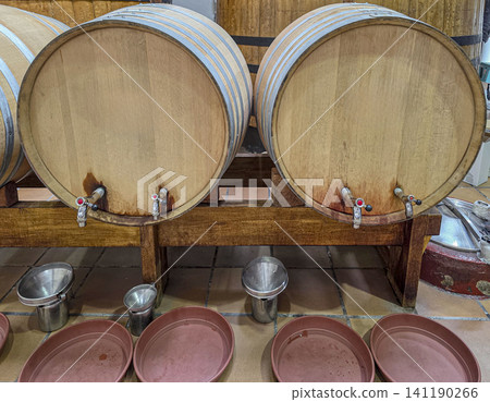 Wooden oak wine barrels sit on a rustic stand in a cellar. Metal taps are ready to pour wine. Plates and cups are placed nearby for tasting. The scene shows the process of wine production. Wooden oak wine barrels sit on a rustic stand in a cellar. Metal taps are ready to pour wine. Plates and cups are placed nearby for tasting. The scene shows the process of wine production. 141190266