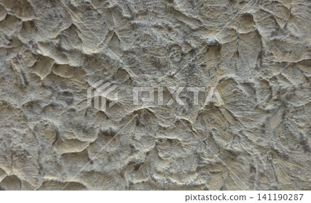 The surface shows a rough grey stucco wall with uneven texture. The decorative finish adds depth and interest to the plaster material that covers the wall. The surface shows a rough grey stucco wall with uneven texture. The decorative finish adds depth and interest to the plaster material that covers the wall. 141190287