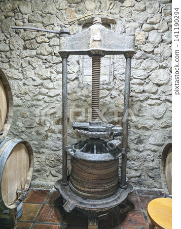 An ancient grape press is located in a rustic wine cellar with stone walls. Wooden barrels are nearby, holding wine. This scene shows the process of winemaking from the past. An ancient grape press is located in a rustic wine cellar with stone walls. Wooden barrels are nearby, holding wine. This scene shows the process of winemaking from the past. 141190294