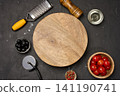 A round empty wooden board on a dark background with tomatoes, olives, spices, oil, a grater, and a knife. 141190741