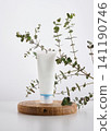 An empty white cosmetic tube stands on a round wooden podium surrounded by eucalyptus branches 141190746