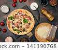Appetizing round pizza on a black board surrounded by cherry tomatoes, olives, grated cheese, spices and kitchen utensils 141190768