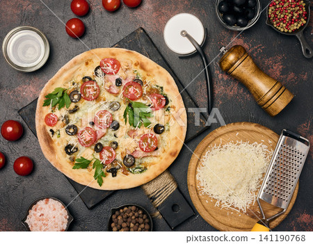 Appetizing round pizza on a black board surrounded by cherry tomatoes, olives, grated cheese, spices and kitchen utensils Appetizing round pizza on a black board surrounded by cherry tomatoes, olives, grated cheese, spices and kitchen utensils 141190768
