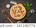 Whole pizza on a wooden board surrounded by tomatoes, olives, herbs, spices and kitchen tools on a dark background 141190774