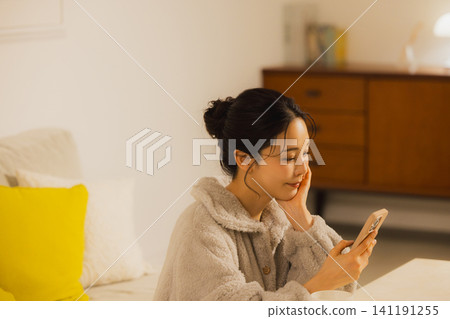 一個年輕的女人,看著她的智能手機在客廳裡在晚上 一個年輕的女人,看著她的智能手機在客廳裡在晚上 141191255