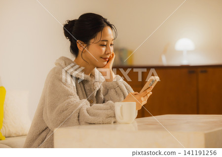 一個年輕的女人,看著她的智能手機在客廳裡在晚上 一個年輕的女人,看著她的智能手機在客廳裡在晚上 141191256