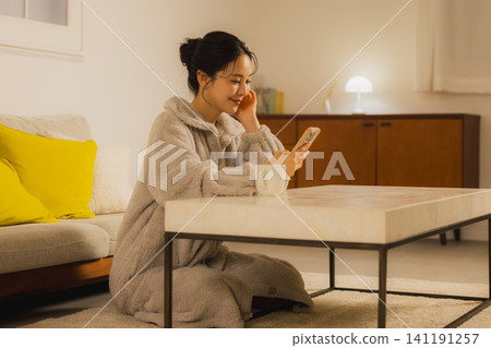一個年輕的女人,看著她的智能手機在客廳裡在晚上 一個年輕的女人,看著她的智能手機在客廳裡在晚上 141191257