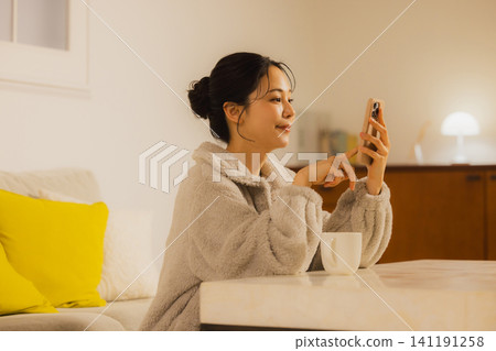 一個年輕的女人，看著她的智能手機在客廳裡在晚上 141191258