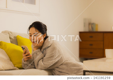一個年輕的女人,看著她的智能手機在客廳裡在晚上 一個年輕的女人,看著她的智能手機在客廳裡在晚上 141191259