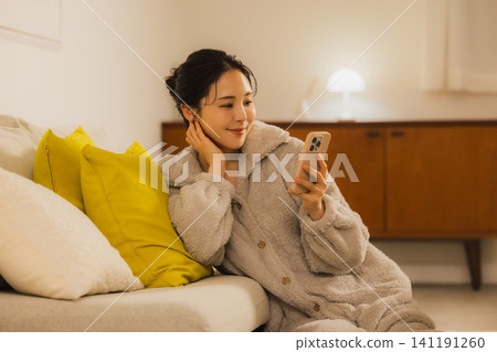 一個年輕的女人,看著她的智能手機在客廳裡在晚上 一個年輕的女人,看著她的智能手機在客廳裡在晚上 141191260
