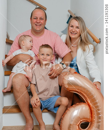 a heartwarming family portrait of a man holding a baby girl, a woman sitting next to a boy, and the boy in front of the man, all sitting together on a wooden staircase with metal handrails. a heartwarming family portrait of a man holding a baby girl, a woman sitting next to a boy, and the boy in front of the man, all sitting together on a wooden staircase with metal handrails. 141191308