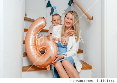 A heartwarming family portrait of a mother and son celebrating a birthday on a wooden staircase. The scene is set indoors with a blue balloon and a white hat, capturing the celebratory mood. A heartwarming family portrait of a mother and son celebrating a birthday on a wooden staircase. The scene is set indoors with a blue balloon and a white hat, capturing the celebratory mood. 141191335