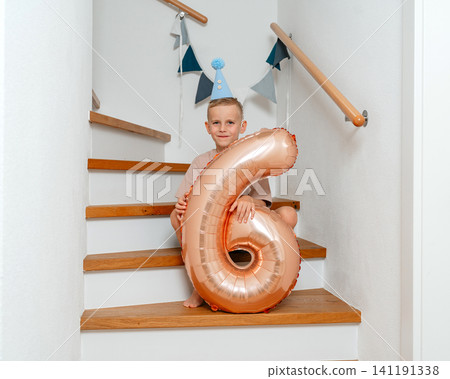 A boy wearing a party hat holds balloon number six on a wooden staircase adorned with wall decorations, capturing a celebratory front view of a children's birthday party with blue and white. A boy wearing a party hat holds balloon number six on a wooden staircase adorned with wall decorations, capturing a celebratory front view of a children's birthday party with blue and white. 141191338