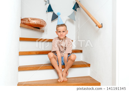 Boy sitting on stairs, in indoor staircase with white walls and wooden handrail, with staircase, wooden handrail, party hat, balloon, with wood, plastic texture, from a frontal view perspective. Boy sitting on stairs, in indoor staircase with white walls and wooden handrail, with staircase, wooden handrail, party hat, balloon, with wood, plastic texture, from a frontal view perspective. 141191345