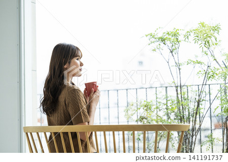 A young woman drinking coffee by the window on a rainy day. 141191737