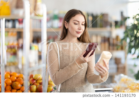 Smiling young girl shopper carefully examining mayonnaise on shelves of food store, scan QR code Smiling young girl shopper carefully examining mayonnaise on shelves of food store, scan QR code 141193170