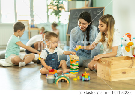 Children playing in living room of kindergarten, babysitter support and participate in game. Children playing in living room of kindergarten, babysitter support and participate in game. 141193282
